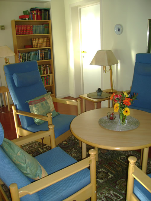 Bilde av en stue med blå lenestoler som står rundt et rundt trebord med en bokhylle i bakgrunnen